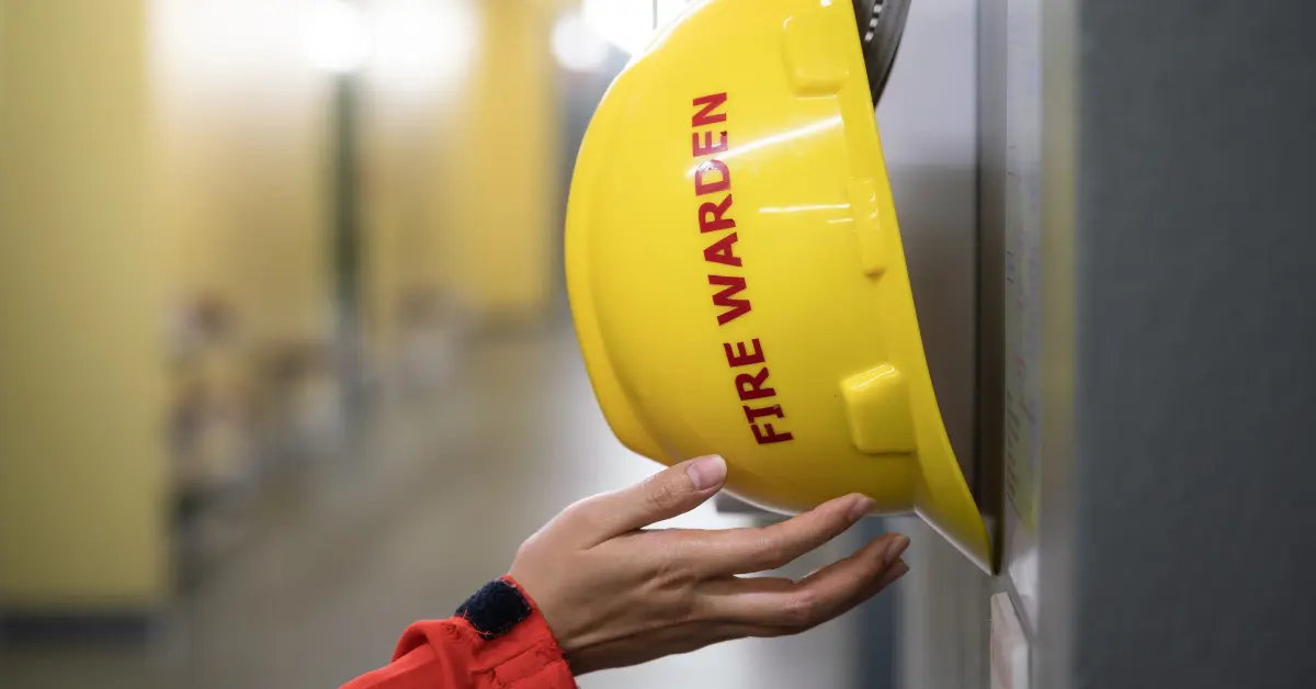 Fire warden yellow hard hat hanging on a wall with woman's hand underneath